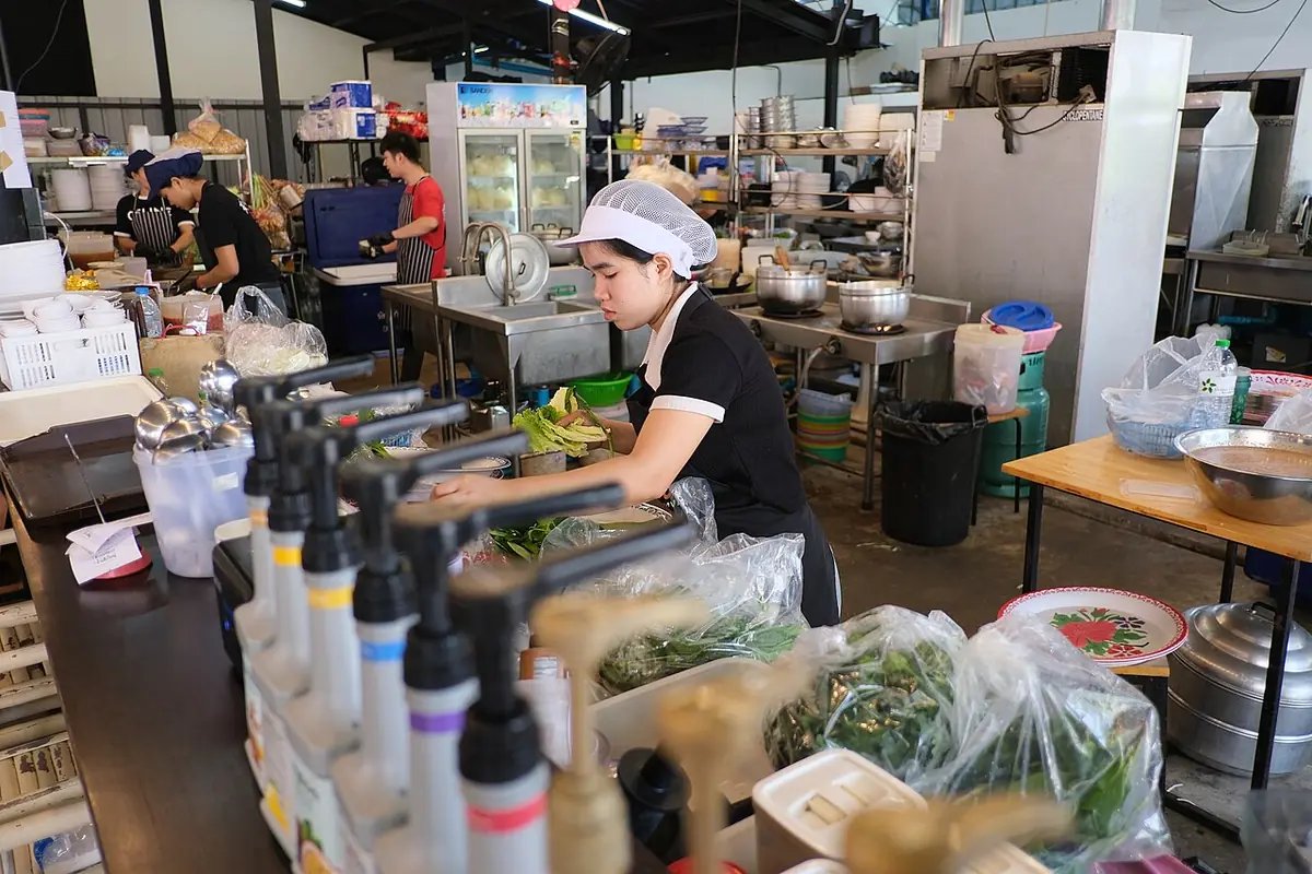 A cook wearing a hairnet prepares fresh greens at a stainless-steel counter in a commercial kitchen